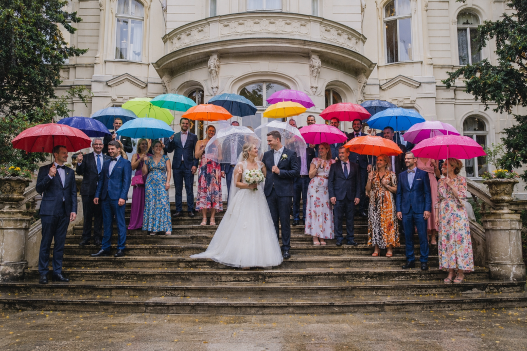 Hochzeitsgesellschaft mit bunten Regenschirmen auf einer Schlosstreppe, Brautpaar unter durchsichtigem Schirm bei Regen